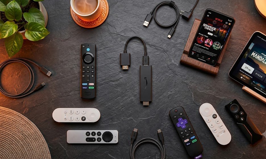 Top-down flat lay of streaming devices including Amazon Fire TV Stick, remote controls, smartphone, and tablet arranged on a dark surface with warm orange accent lighting